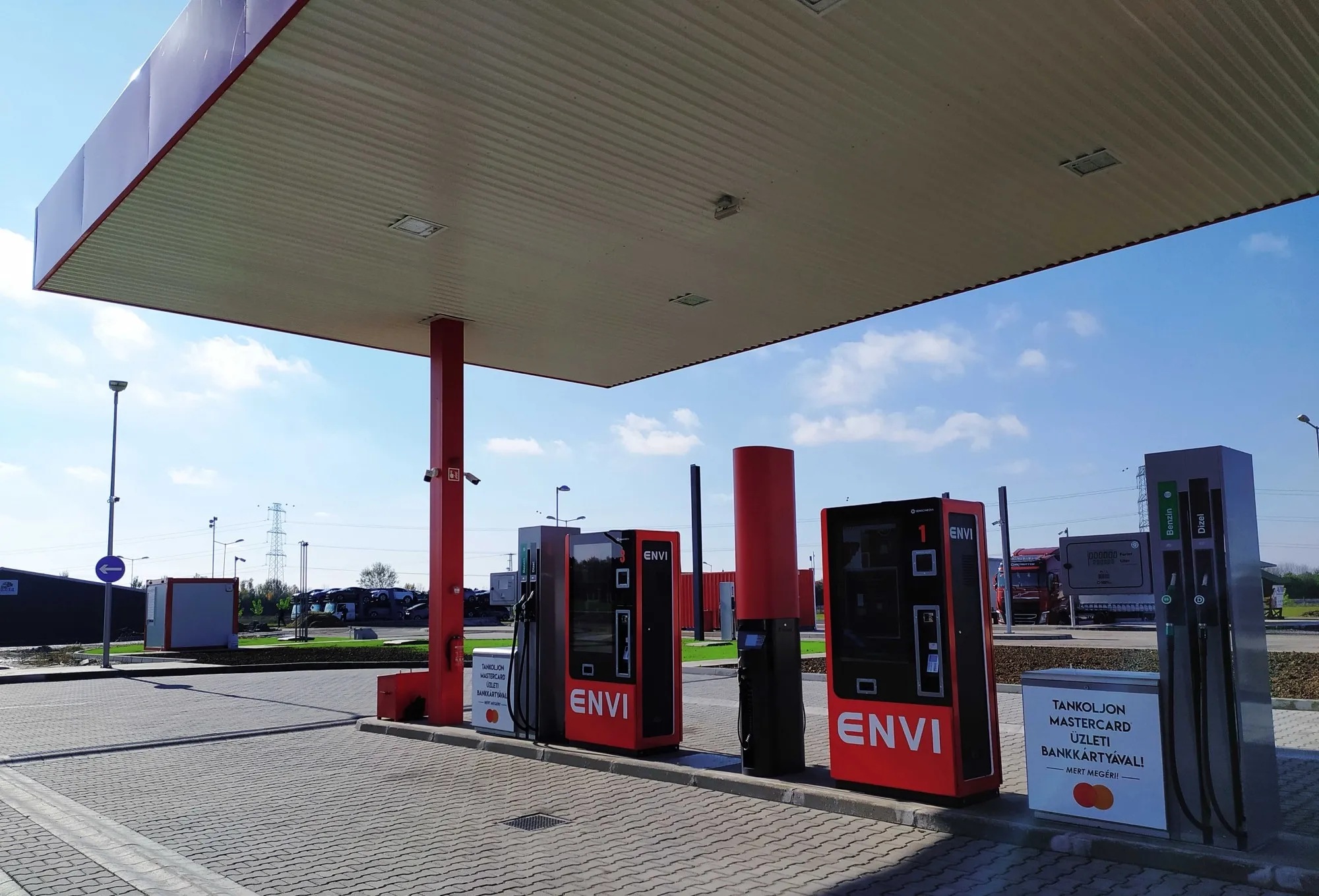 ENVI töltőállomás piros oszlopokkal és fizetőautomatákkal, kék ég alatt